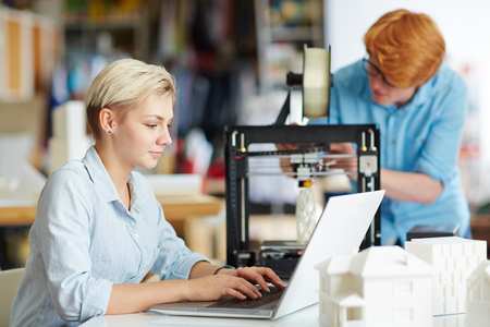 Young designer networking on background of her colleague printing architectural modelの写真素材