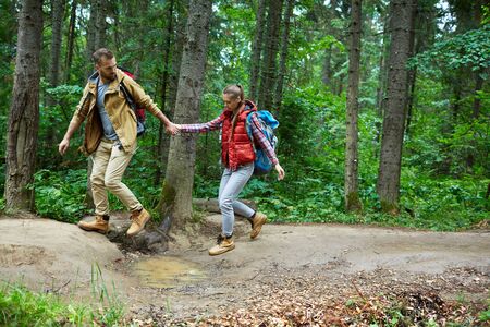 Young tourist helping his girlfriend pass over puddle in forest