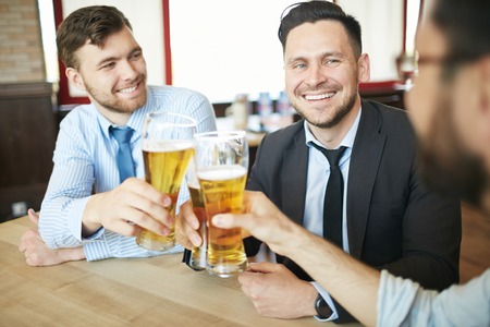 Three laughing businessmen in formal clothes having beer in bar after work celebrating with clinking glassesの写真素材