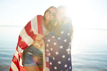 Two pretty girls wrapped in national American flag looking at camera on sunny day