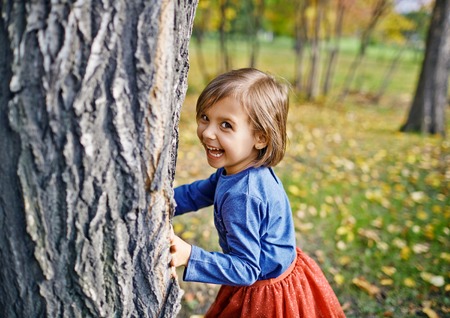 Cute little girl hiding behind tree in autumn park, looking sideways at camera laughing as she tries...