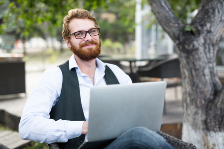 Happy young bearded man with laptop networking outdoorsの写真素材