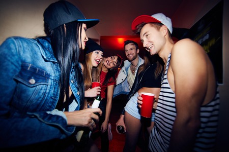 Low angle group of young stylish people standing in dark hall of nightclub together, talking, laughing and drinking beer as they wait to be admittedの写真素材