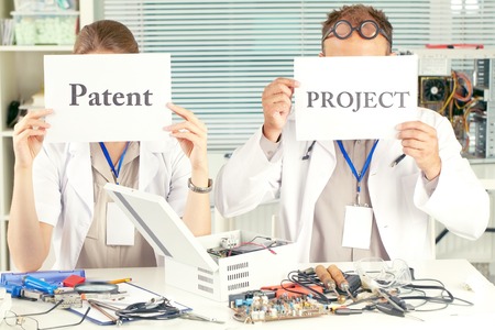 Scientists sitting at the table and holding placard with the words patent and project in front of their facesの写真素材