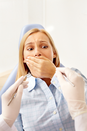 A young woman in dentist chair covering her mouth before dental toolsの写真素材