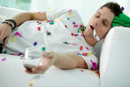 Young man sleeping on sofa in party hat all in confetti and with a wine glassの写真素材