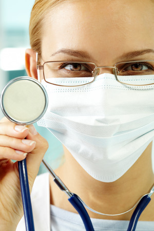 Portrait of a young doctor in mask and with stethoscope looking at camera and smilingの写真素材