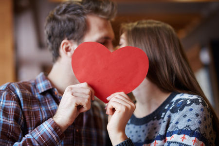 Affectionate valentines kissing behind red paper heartの写真素材