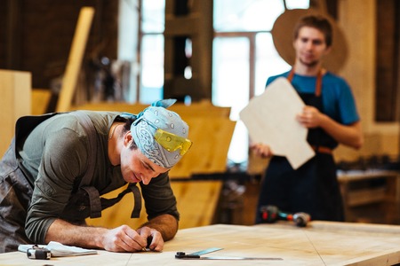 Joiner or carpenter taking measures with his assistant on background
