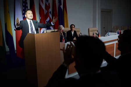 Formally-dressed middle-aged man standing on stage, giving public emotional speech, gesticulating in conference room on background of national flagsの写真素材