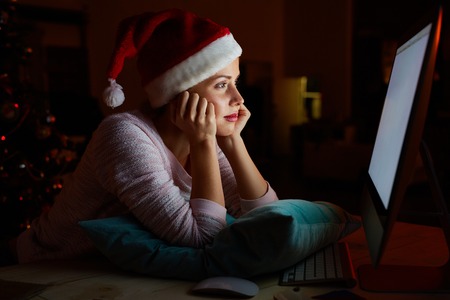 Bored girl in Santa cap looking at computer monitor on xmas nightの写真素材