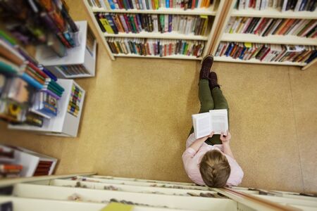 Teenage girl sitting in library aisle between bookshelves and reading interesting book