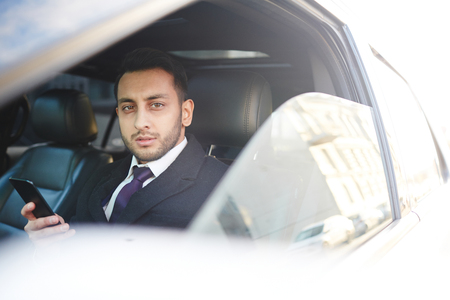 Handsome young businessman with smartphone sitting in his car and looking at cameraの写真素材