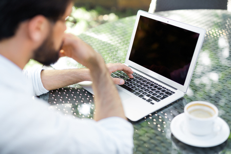 Bearded financial manager with rolled up shirt sleeves searching mistake in his calculations on laptop while drinking coffee in outdoor cafe, over shoulder viewの写真素材