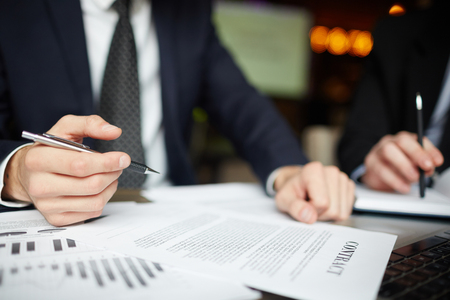 Closeup portrait of two unrecognizable business people wearing black formal suits signing contract papers at table during meetingの写真素材