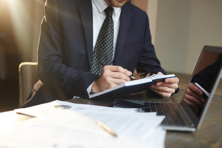 Closeup portrait of unrecognizable successful businessman wearing black formal suit  writing in planner while working with documentation and laptopの写真素材