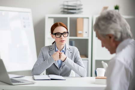 Young employee sitting by desk and making report to her director or sharing her business ideasの写真素材