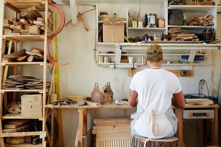 Back view of young contemporary artisan sitting by workplace on stump seat