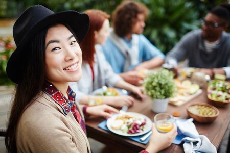 Happy young Asian female in hat looking at camera while sitting by festive table with her friendsの写真素材