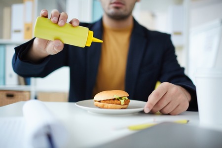 Office manager adding mustard from plastic bottle to his hamburger before eating itの写真素材