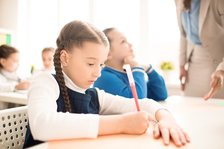 Serious schoolgirl doing exercise at the table at classroomの写真素材