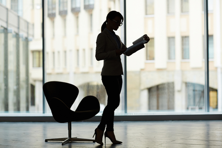 Profile view of unrecognizable manager standing at office room with panoramic windows and studying document with concentrationの写真素材