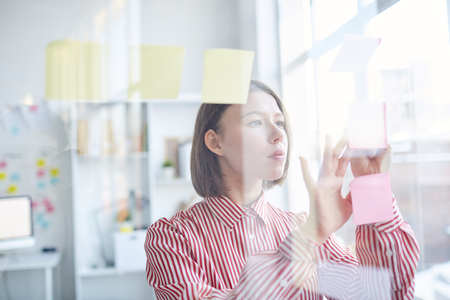 Young attractive caucasian woman in striped blouse reading notes on glass wall in modern office