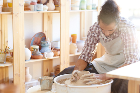 Young professional African American craftsman sitting in workshop and making clay ware on pottery wheelの写真素材