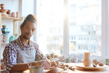 Portrait of a handsome confident mixed race man in apron looking at camera while making clay vases in workshopの写真素材