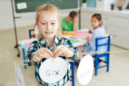 Cute blond primary schoolgirl playing math game while having indoor activities with classmates after lessonsの写真素材