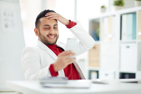 Smiling businessman sitting at his workplace and using smartphoneの写真素材