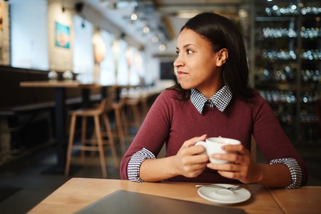 Portrait of a beautiful mixed race woman sitting in a cup of coffee and thoughtfully looking at table in restaurantの写真素材