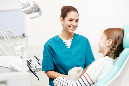 Positive child beautiful young female pediatric dentist in uniform talking on a laptopの写真素材