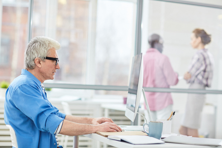 Side view of mature gray-haired Caucasian man in glasses sitting at desk and working on computer in modern officeの写真素材