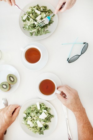 Overview of two cups of tea, sunglasses and two kiwi halves on table while humans eating fresh vegetarian saladの写真素材