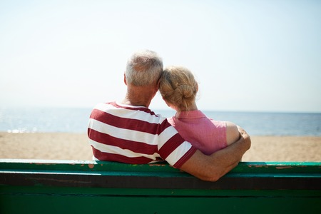 Back view of senior affectionate couple sitting on bench by waterside on hot summer dayの写真素材