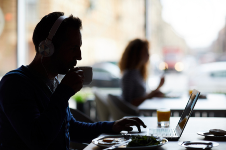 Silhouette of male hipster entrepreneur in headphones sitting at table and using laptop while drinking coffee and working in cafeの写真素材