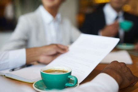 Close-up of unrecognizable advisor sitting at table with coffee cup and examining contract while...