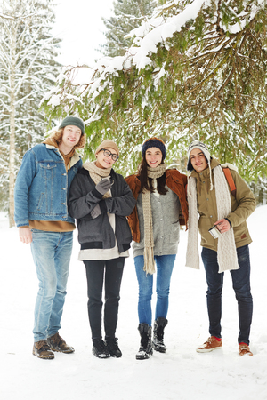 Full length portrait of group of happy young people posing in winter resort standing under fir tree in beautiful snowy forestの写真素材