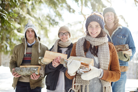 Waist up portrait of group of happy young people posing in beautiful winter forest holding logs and...