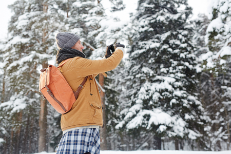 Positive curious young man in winter coat wearing satchel using smartphone while photographing nature during hiking, travel conceptの写真素材