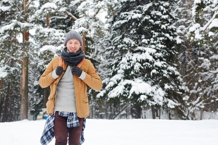 Smiling handsome young guy in knitted hat and scarf carrying backpack going down hill while hiking in winter forestの写真素材