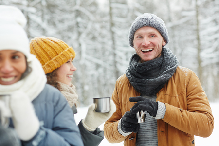 Cheerful excited young friends in warm clothing standing in winter forest and drinking hot tea to get warm while chatting together during hikingの写真素材