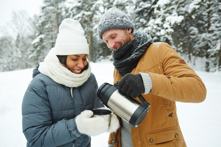 Cheerful careful young man in snowy hat and scarf pouring hot tea from 
to girlfriend while she holding mug in winter forestの写真素材