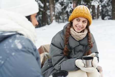 Positive pretty girl in yellow knitted hat talking to friend and drinking tea from mug at camping in winter forestの写真素材