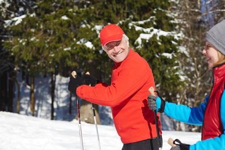 Happy senior sportsman looking at his spouse while both moving on skis or trekking in winter forestの写真素材