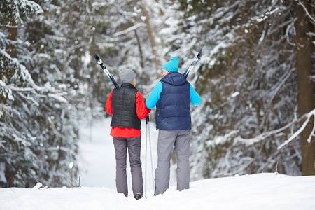 Rear view of sporty aged couple with skiing equipment walking down forest path on winter dayの写真素材