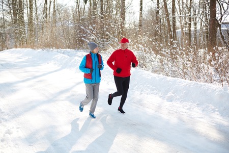 Aged sporty couple having talk while running on snow road on sunny winter dayの写真素材