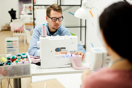 Serious concentrated young man in glasses sitting at table and sewing garment while working on sewing machine in tailoring studioの写真素材