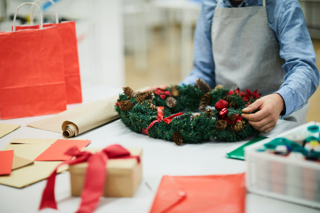 Close-up of unrecognizable man in apron standing at table with papers and making holiday wreath while preparing Christmas decoration in workshopの写真素材